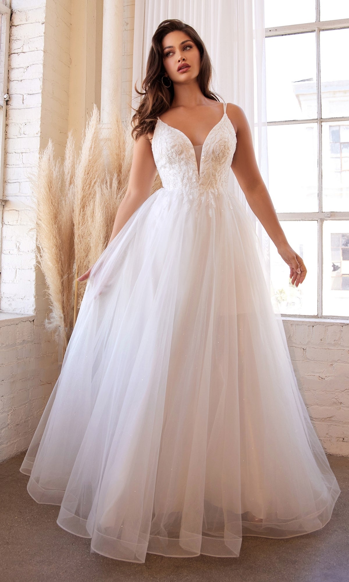A woman poses indoors near large windows and pampas grass, showcasing the Off-White Long A-Line Wedding Dress CD0154W with a deep V-neckline and tulle skirt—an elegant and affordable bridal gown.